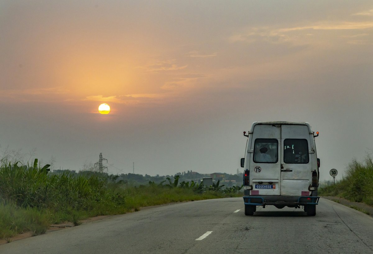Minibus blanc roulant sur une route bordée de végétation au coucher du soleil, près d'Abidjan